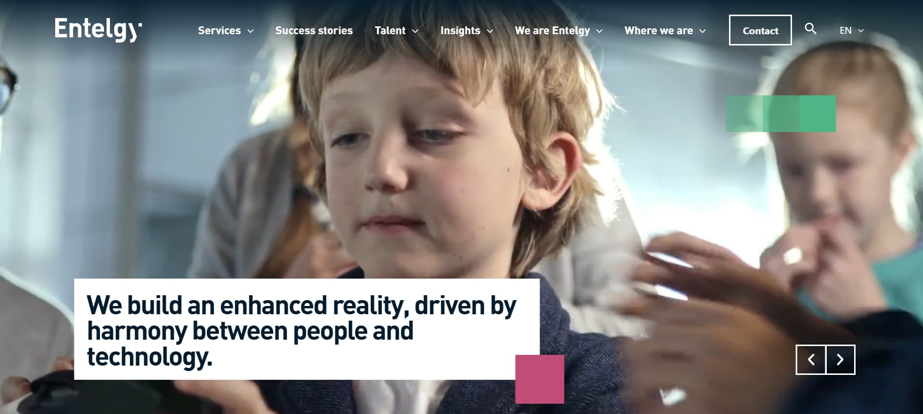 Corporate website banner for Entelgy showing a young child in a classroom or learning environment with adults and another child blurred in the background, overlaid with the text “We build an enhanced reality, driven by harmony between people and technology.”
