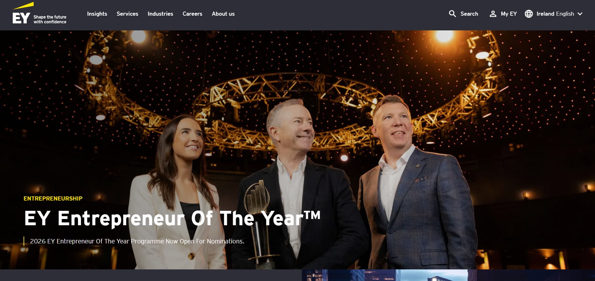 Event stage photograph with three business professionals in formal attire under circular stage lighting, promoting “EY Entrepreneur Of The Year” with nomination information.